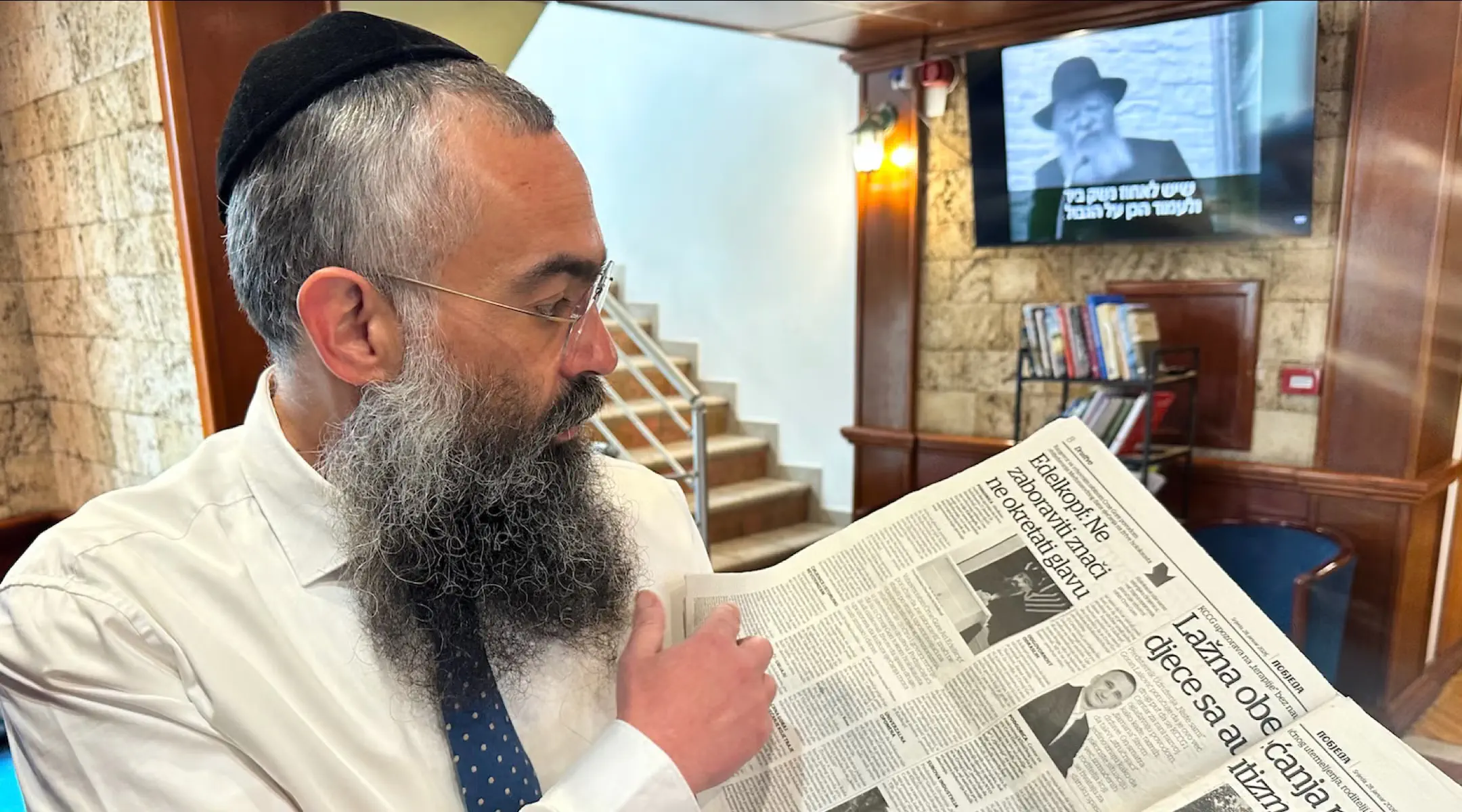 Rabbi Ari Edelkopf, a Los Angeles-born Chabad emissary, reads a story about himself in a Podgorica newspaper in April 2026. Edelkopf heads the Religious Jewish Community of Montenegro. Credit: Larry Luxner